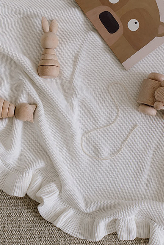 White knitted baby blanket with frilled edge on the floor with wooden children's toys