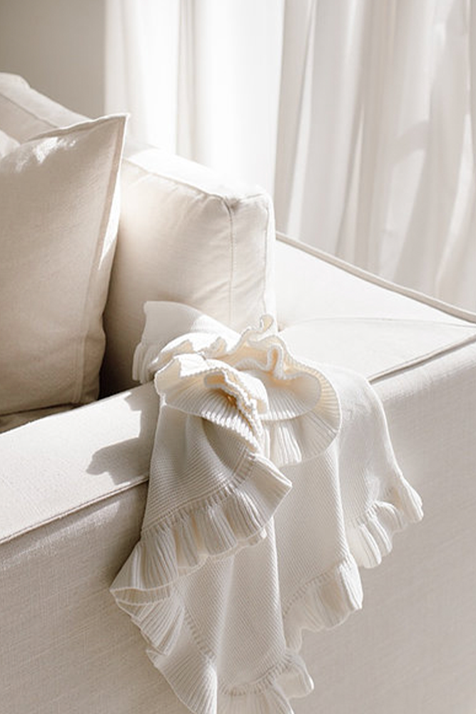 White knitted baby blanket with frilled edge on a white couch with sheer curtains in the background and morning light.
